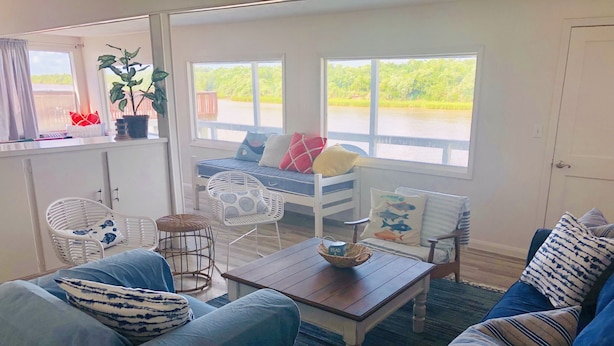 A cozy living room with a sofa, armchair, and a small table with a basket. There are two windows with views of greenery and a wooden deck.