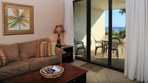 Een woonkamer met een bank, een bijzettafeltje met een mand, een lamp en een schuifdeur naar een terras buiten met stoelen en uitzicht op zee.