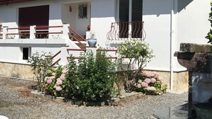 Ein weißes Gebäude mit rotem Ziegeldach, einem Balkon mit Metallgeländer und einem kleinen Garten mit rosa Blumen.