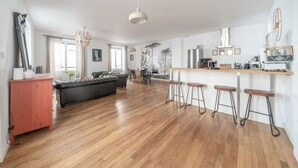 Zona d'estar àmplia amb terra de fusta, un sofà negre, un armari vermell i una zona de cuina amb barra i tamborets.