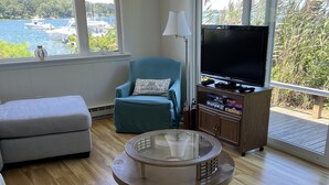 Een woonkamer met een ronde glazen salontafel, een blauwgroene fauteuil, een grijze bank en een televisie op een houten kast. Grote ramen bieden uitzicht op een waterpartij en groen.