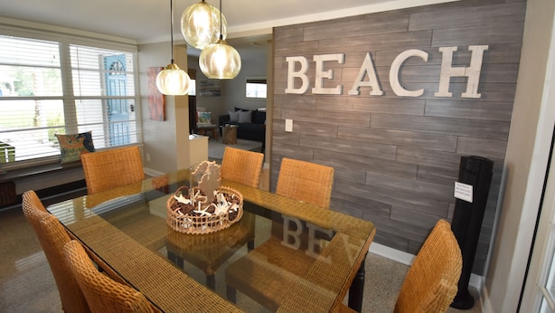 A dining area with a glass table and wicker chairs, a wall with the word "BEACH" in large letters, and a view of the outdoors through the window.