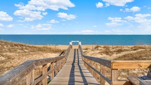 Una pasarela de madera que lleva a una playa, con cielo azul despejado y nubes.