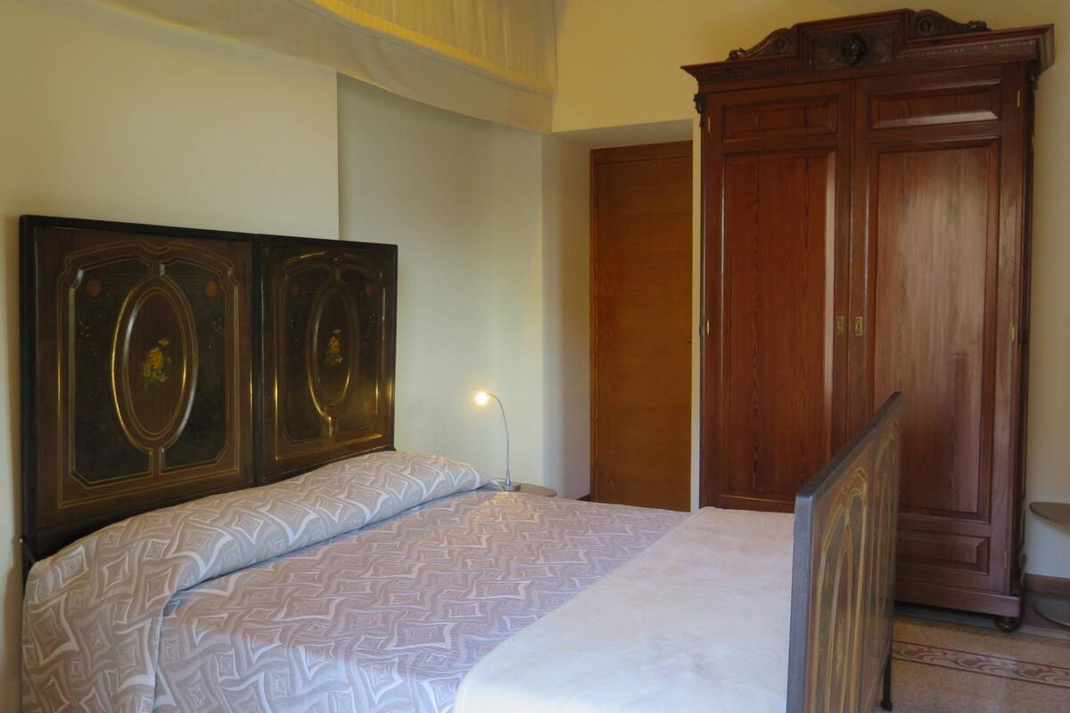 A bedroom with a large bed, a wooden wardrobe, and a nightstand with a lamp.