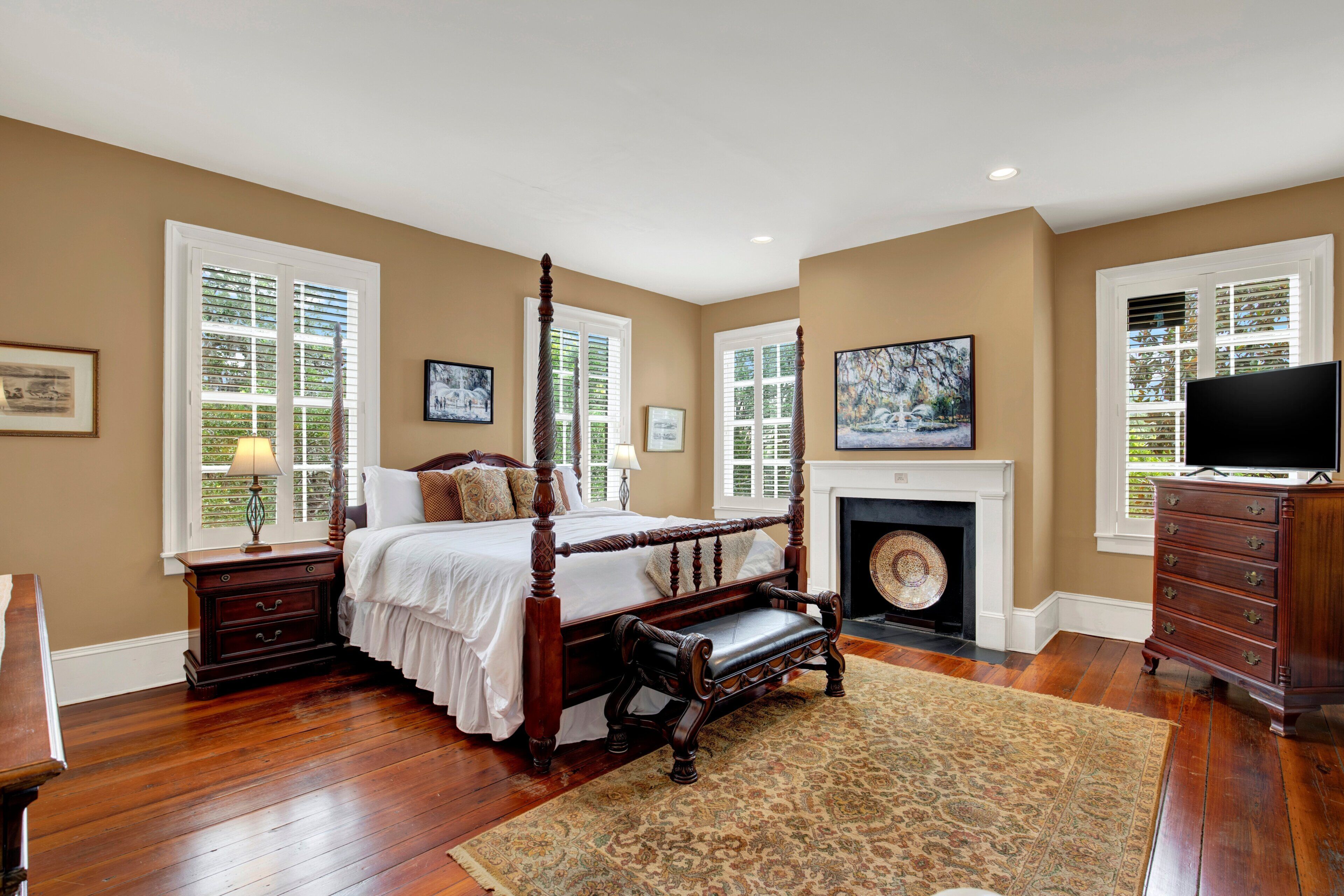 A bedroom with a four-poster bed, a fireplace, a television, and a large window with shutters.