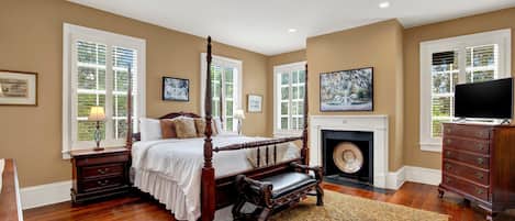 A bedroom with a four-poster bed, a fireplace, a television, and a large window with shutters.