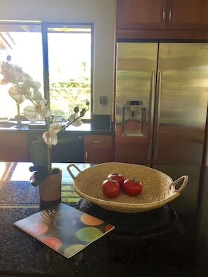 Una cocina con un refrigerador de acero inoxidable, una cesta de mimbre con tomates, un florero con flores blancas y un libro en la encimera.
