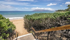 Una playa con una escalera de madera que baja hacia la arena, rodeada de vegetación exuberante.