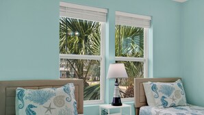 Quarto com duas camas, uma mesa de cabeceira e uma luminária. Janelas com persianas e vista para palmeiras do lado de fora.
