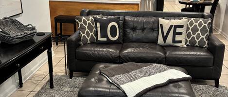A modern living room with a black leather sofa, a matching ottoman, and a patterned armchair. A kitchen with stainless steel appliances is visible in the background.