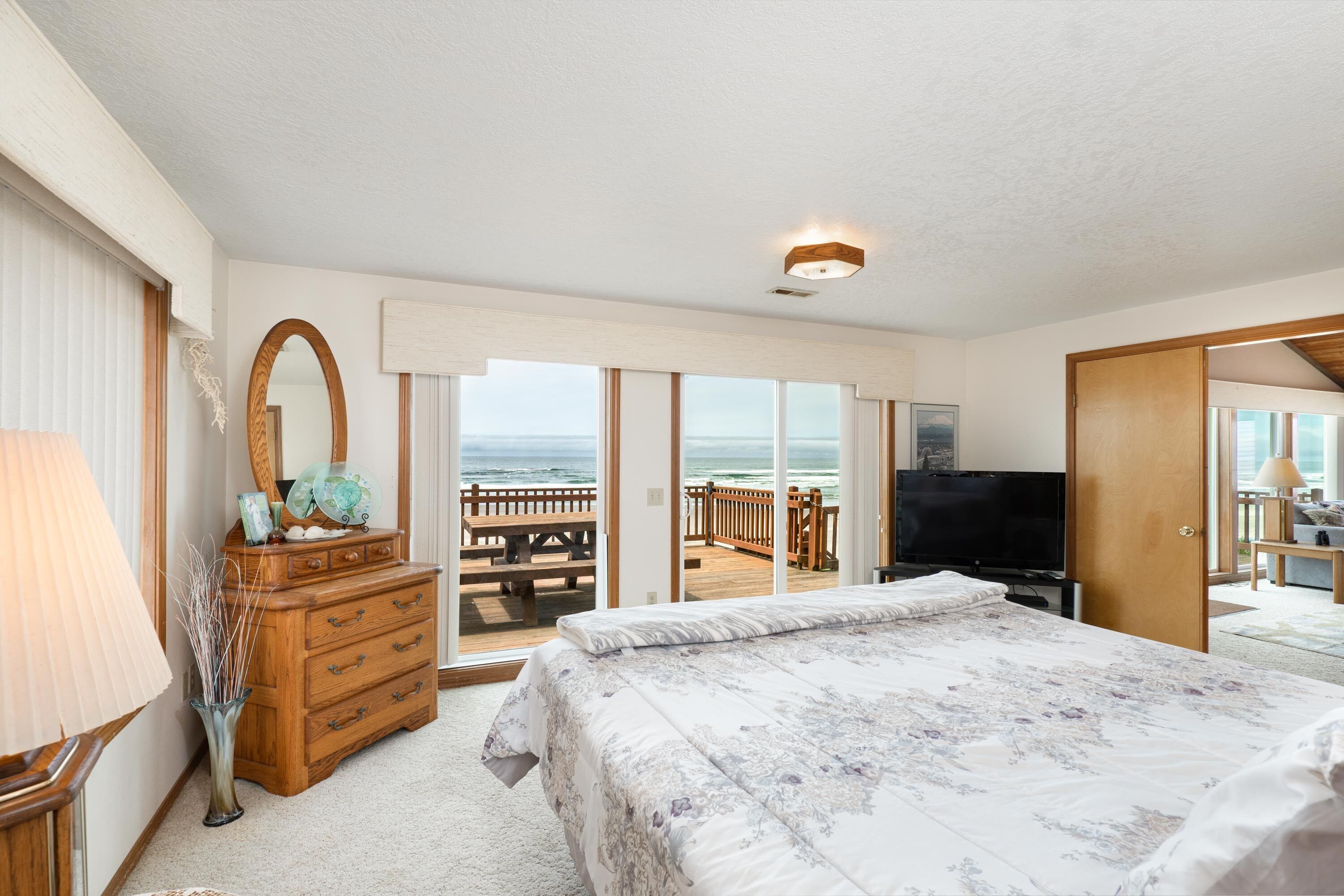 Ein Schlafzimmer mit einem großen Bett, einem Schminktisch, einem Spiegel und einem Fernseher. Es gibt einen Balkon mit Holzterrasse und Meerblick.