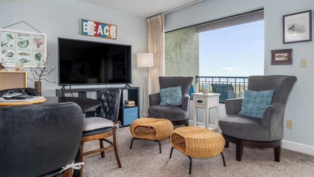 A living room with a flat-screen TV, two grey armchairs with blue cushions, a coffee table, and a wicker ottoman. - Sweeping oceanfront views from this one bedroom condo right on the prom! (Seaside)