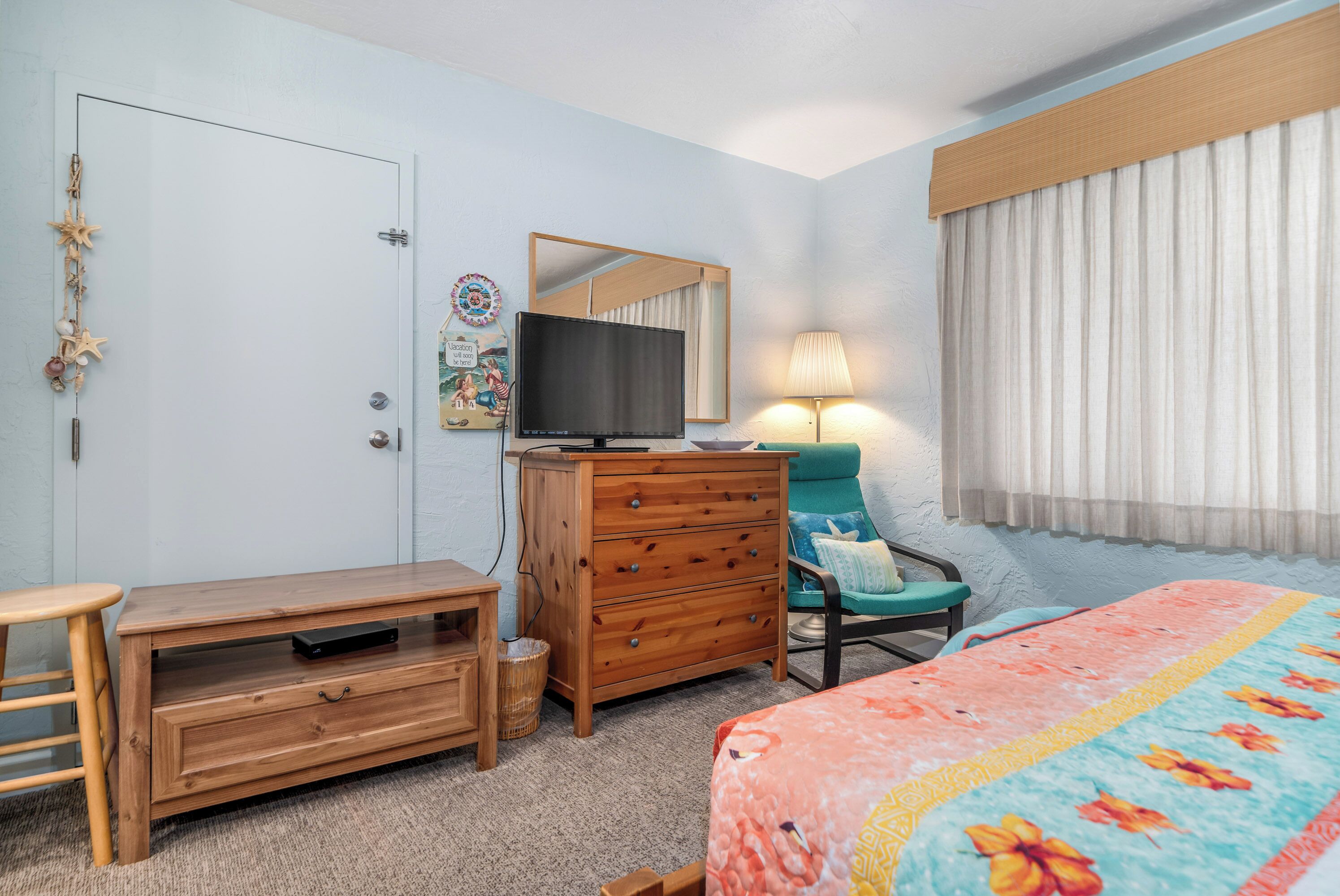 Habitación de hotel con tocador de madera, televisor, una silla, cama con colcha estampada y ventana con cortinas.