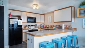 A modern kitchen with a black refrigerator, microwave, and stove. There are blue bar stools and a wooden countertop. - Sweeping oceanfront views from this one bedroom condo right on the prom! (Seaside)