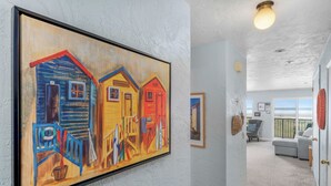 A hallway with a large painting of colorful houses, a light fixture, and a view to a balcony with a chair. - Sweeping oceanfront views from this one bedroom condo right on the prom! (Seaside)
