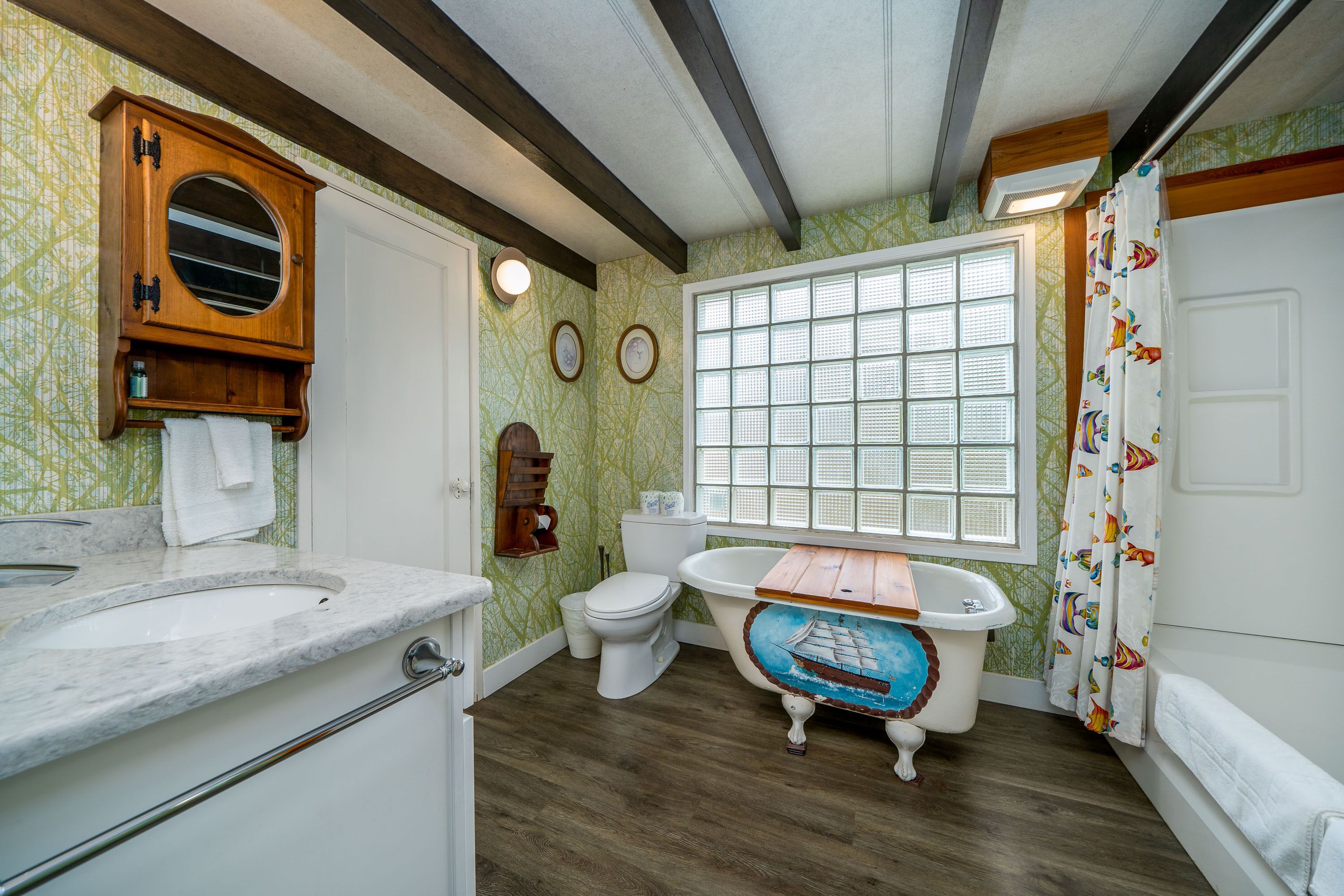 A bathroom with a wooden vanity, a white toilet, a bathtub with a wooden seat, a window with blinds, and a shower curtain with a fish pattern.