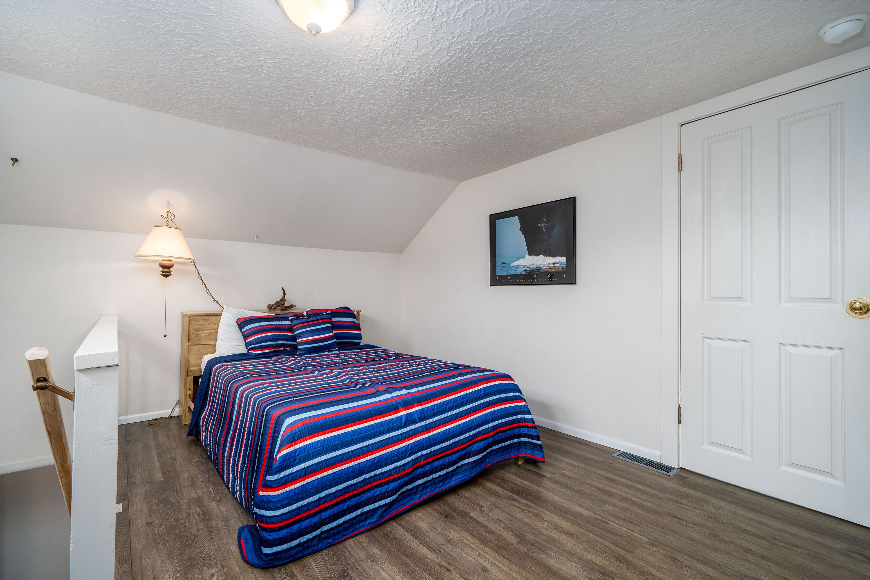 A bedroom with a bed, a nightstand, a lamp, a framed picture on the wall, and a white door.