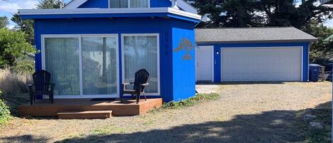 Een klein blauw huis met een garage, twee stoelen buiten en een grindpad.