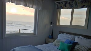 A bedroom with a large window overlooking the ocean, a bed with striped bedding, and a nightstand with a lamp. - Oceanfront Cottage with Deck and Ocean Views, Dogs Ok, Perfect Couples Retreat (Seal Rock)