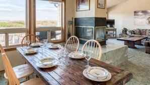Un coin repas avec une table en bois dressée pour quatre personnes, entourée de chaises. On aperçoit une cheminée au fond, et des tableaux encadrés ornent le mur.