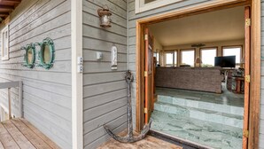Une façade en bois avec un porche, une porte donnant sur l’intérieur et une vue sur l’espace intérieur, où l’on aperçoit un canapé et un téléviseur.