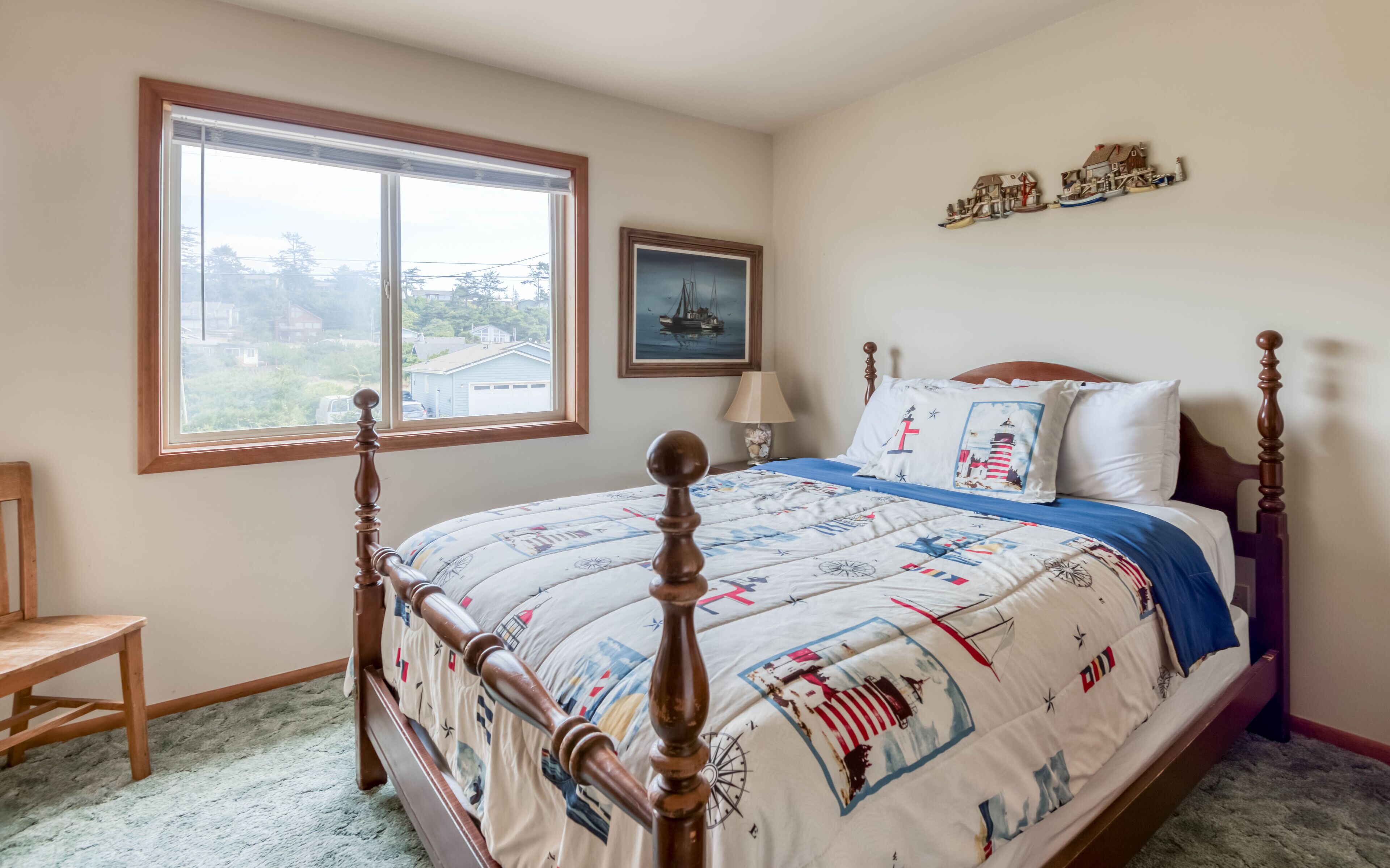 Un dormitorio con un gran ventanal, una cama con cubrecama de temática náutica, cabecera de madera, mesita de noche con lámpara y un cuadro en la pared.