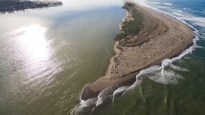 Eine Luftaufnahme einer Küstenlandschaft mit Sandstrand, ruhigem Wasser und sanften Hügeln im Hintergrund.