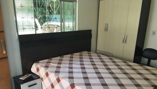 A bedroom with a checkered bedspread, a nightstand with a book, a wardrobe, and a view of the outside through a window.