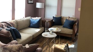 Ein Wohnzimmer mit einem beigefarbenen Sofa, einem gemusterten Sessel, einem kleinen runden Tisch mit Lampe und einem Fenster mit Jalousien.