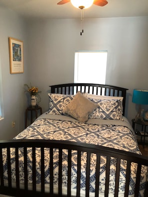 Une chambre à coucher avec un lit, un ventilateur de plafond, une fenêtre, une lampe, une petite table avec un vase et un tableau encadré au mur.