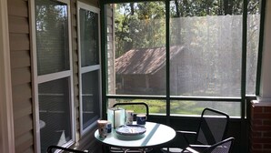Eine überdachte Terrasse mit rundem Tisch und vier Stühlen, einem kleinen Teppich und einem Fenster mit Blick auf ein Haus und Bäume.