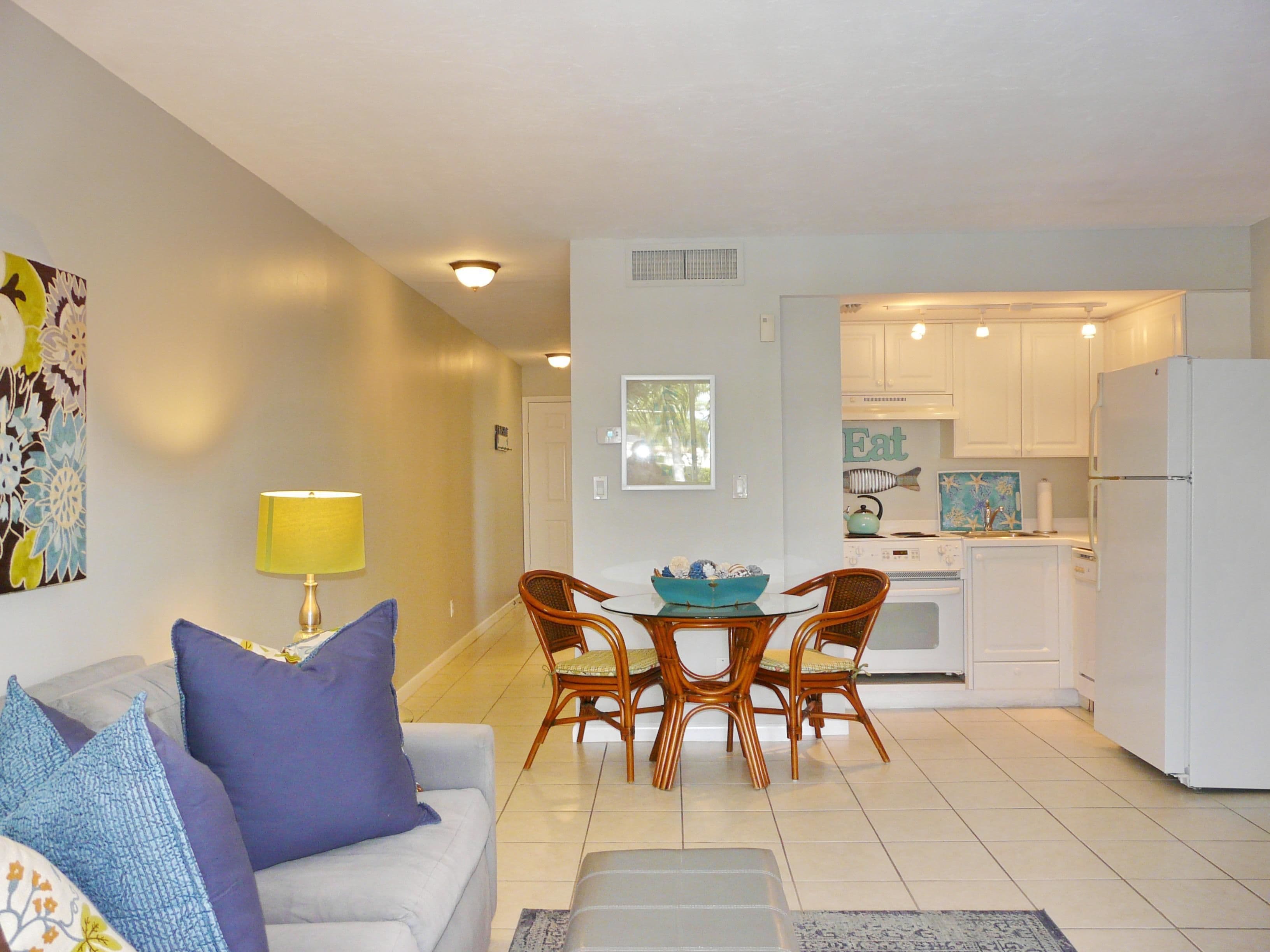 A modern living room with a sofa, a coffee table, and a dining table with chairs. There is a kitchen area with white cabinets and a refrigerator.