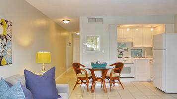 A modern living room with a sofa, a coffee table, and a dining table with chairs. There is a kitchen area with white cabinets and a refrigerator.