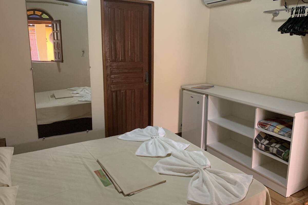 A hotel room with a bed, a wardrobe, a small refrigerator, and a shelf with folded towels.
