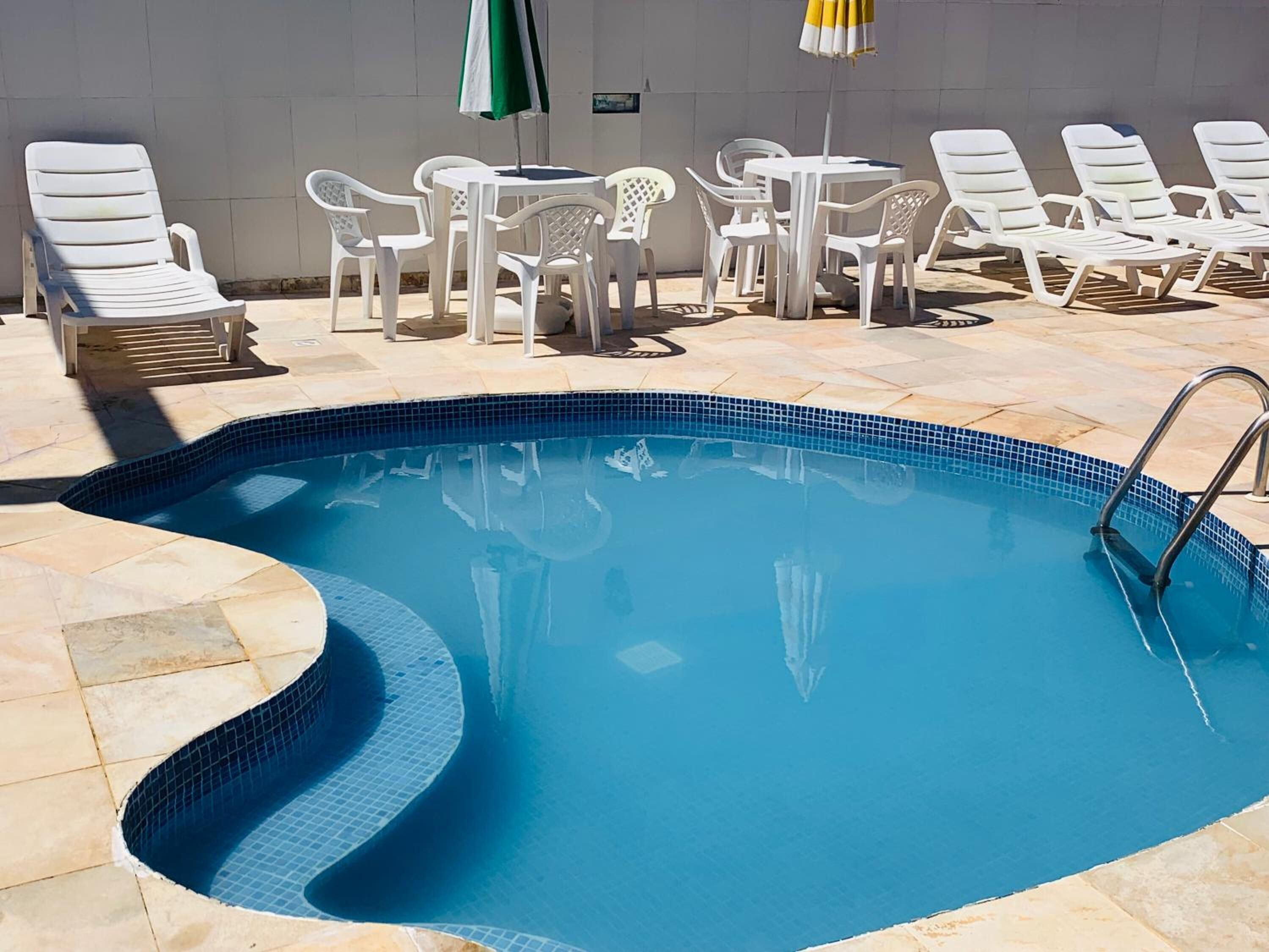 Una piscina con borde de azulejos, rodeada de sillas y mesas de descanso blancas, y un suelo también de azulejos.