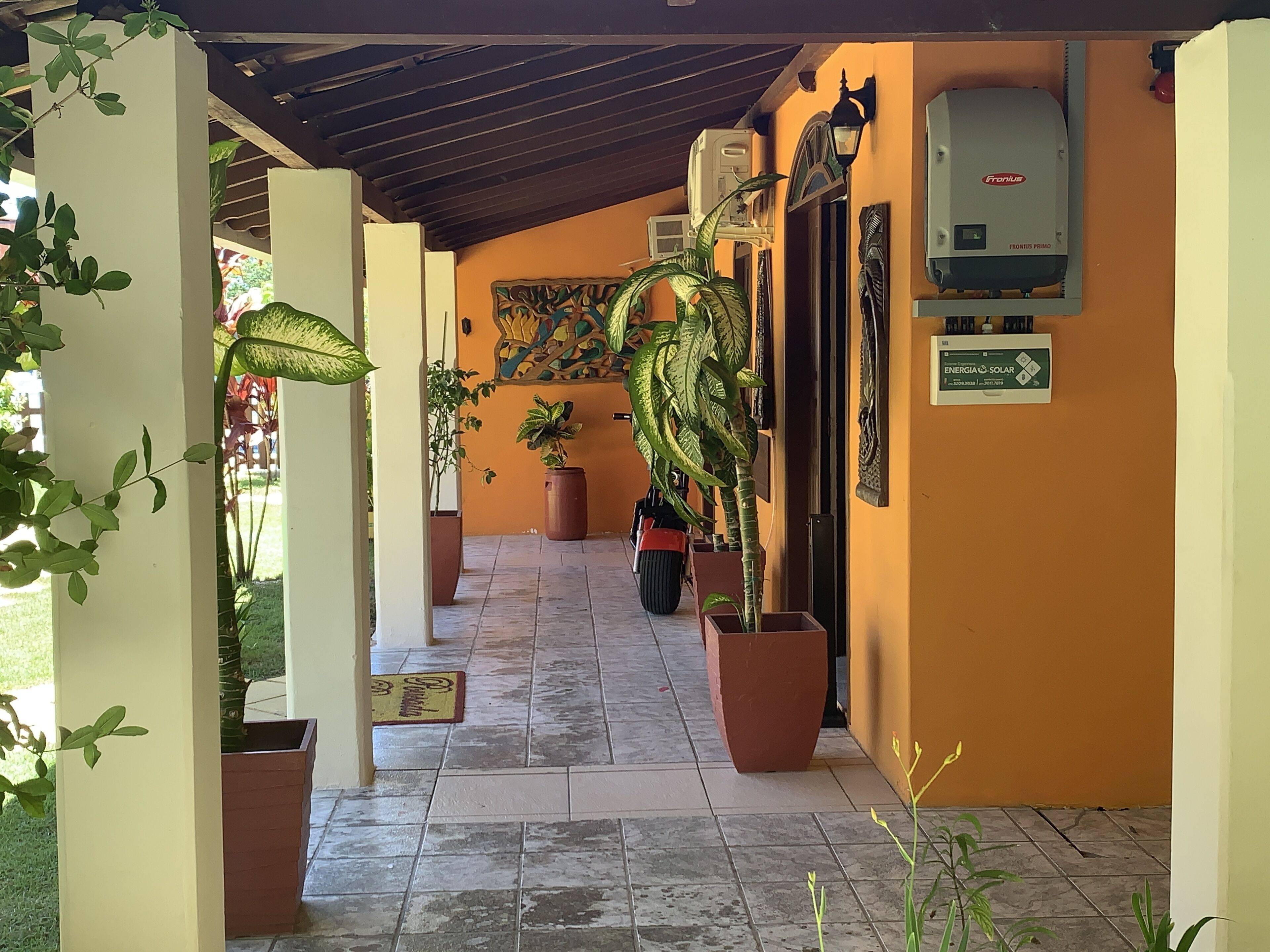 Une allée couverte agrémentée de plantes en pot, un climatiseur fixé au mur et un panneau indiquant l’utilisation d’énergie solaire.