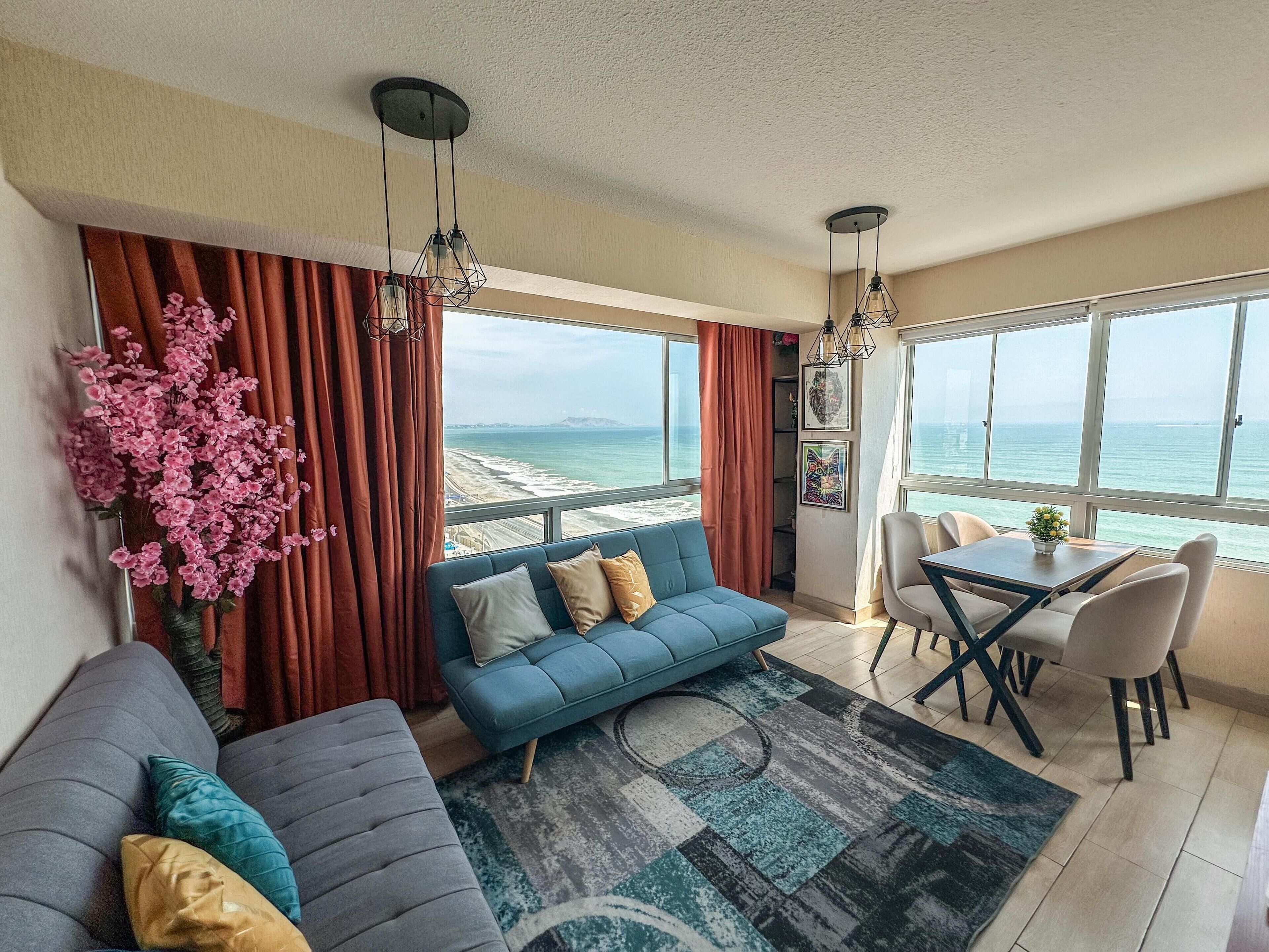 A modern living room with a blue sofa, a dining table with chairs, and a view of the ocean.