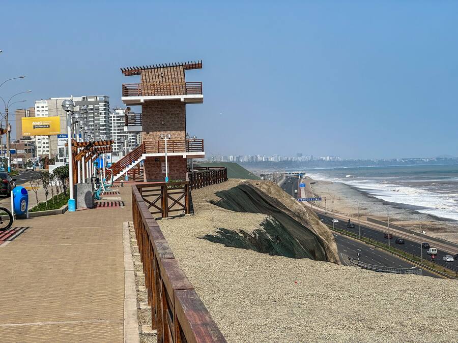 Un paseo marítimo con una pasarela de madera, una playa y un paisaje urbano al fondo.