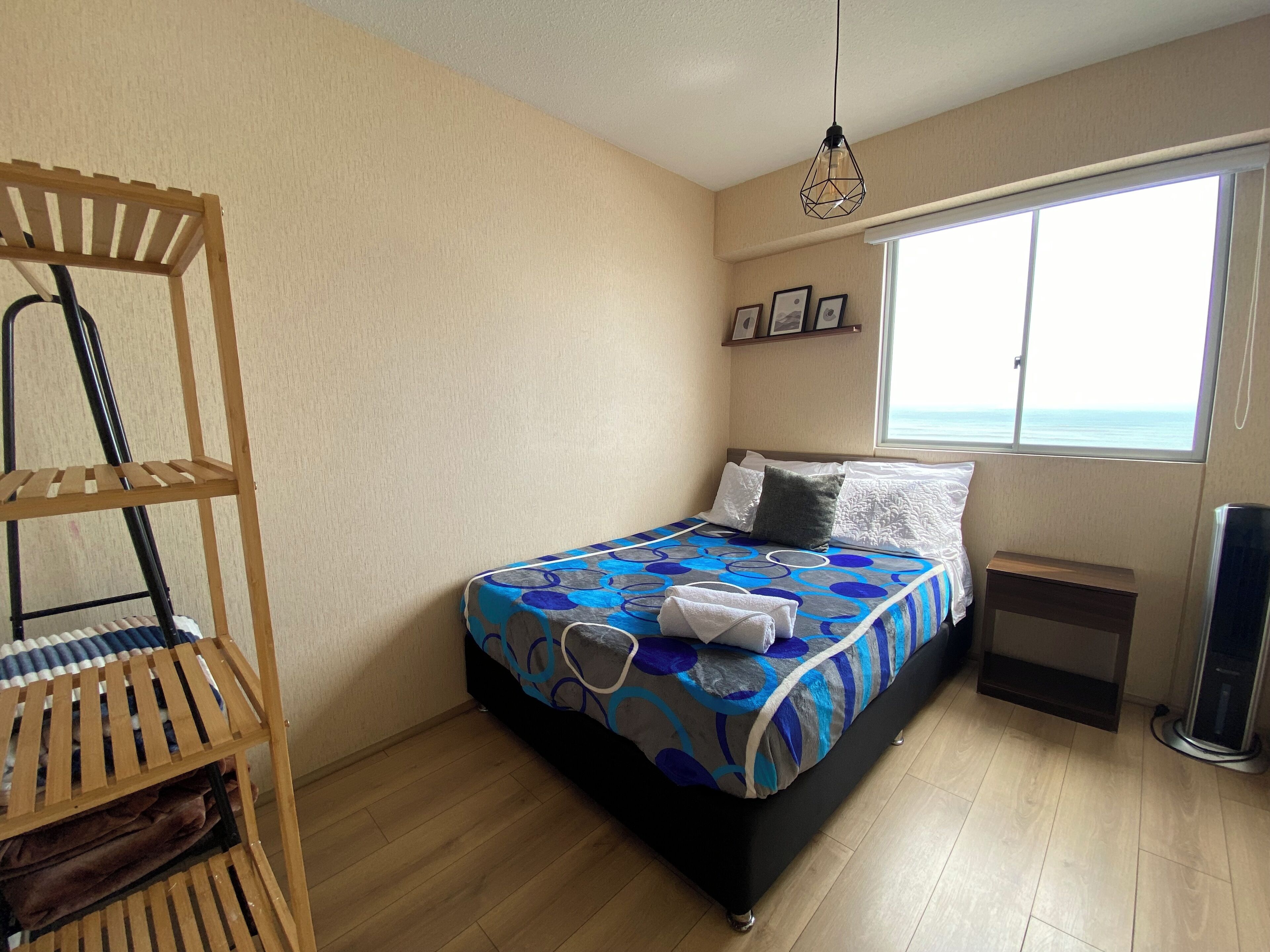 A bedroom with a bed, a wooden shelf, a window with a view, and a ceiling light.