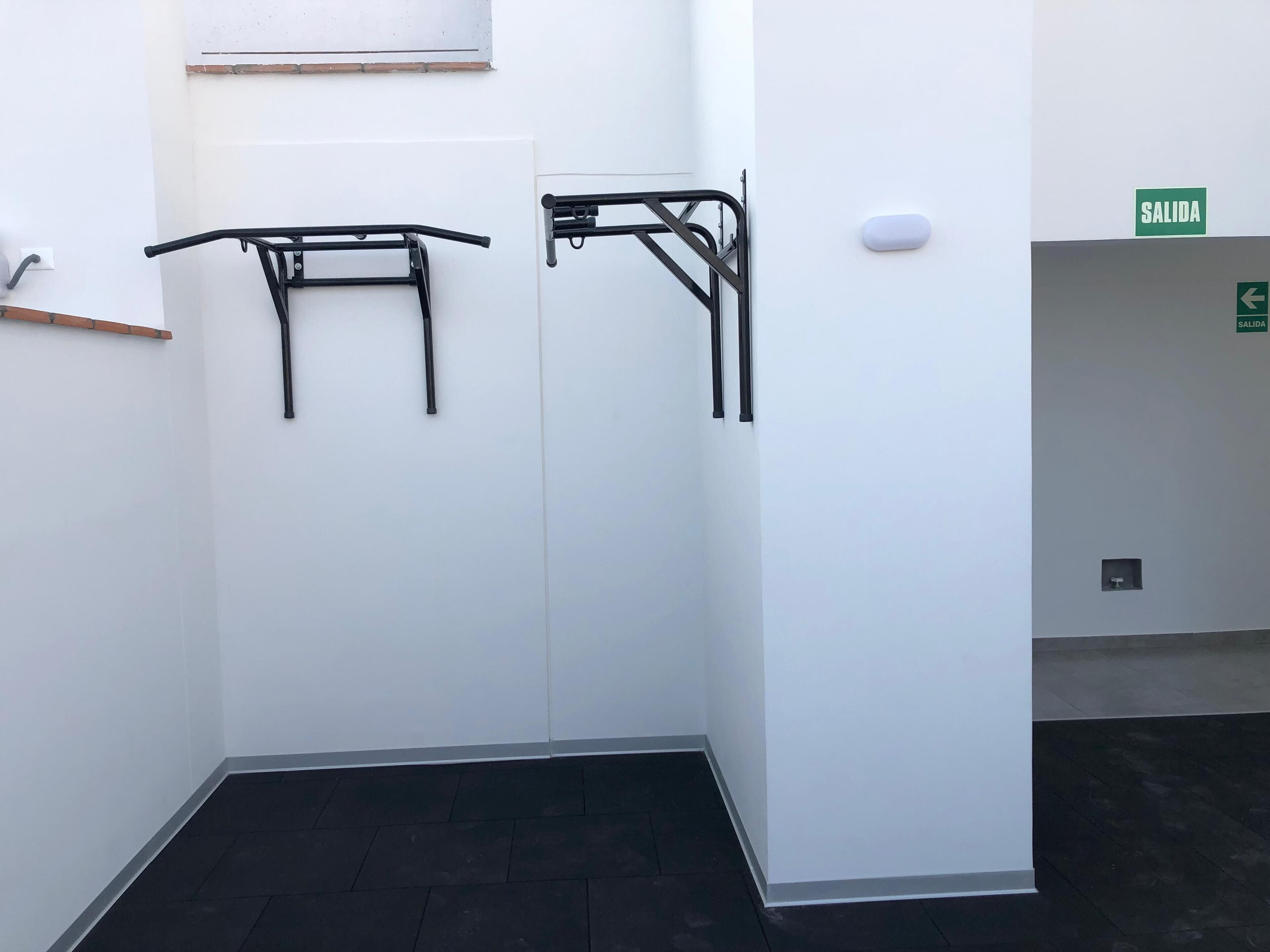 Een smalle gang met twee zwarte metalen leuningen aan witte muren, een groen EXIT bord en een klein raam met houten zonwering.