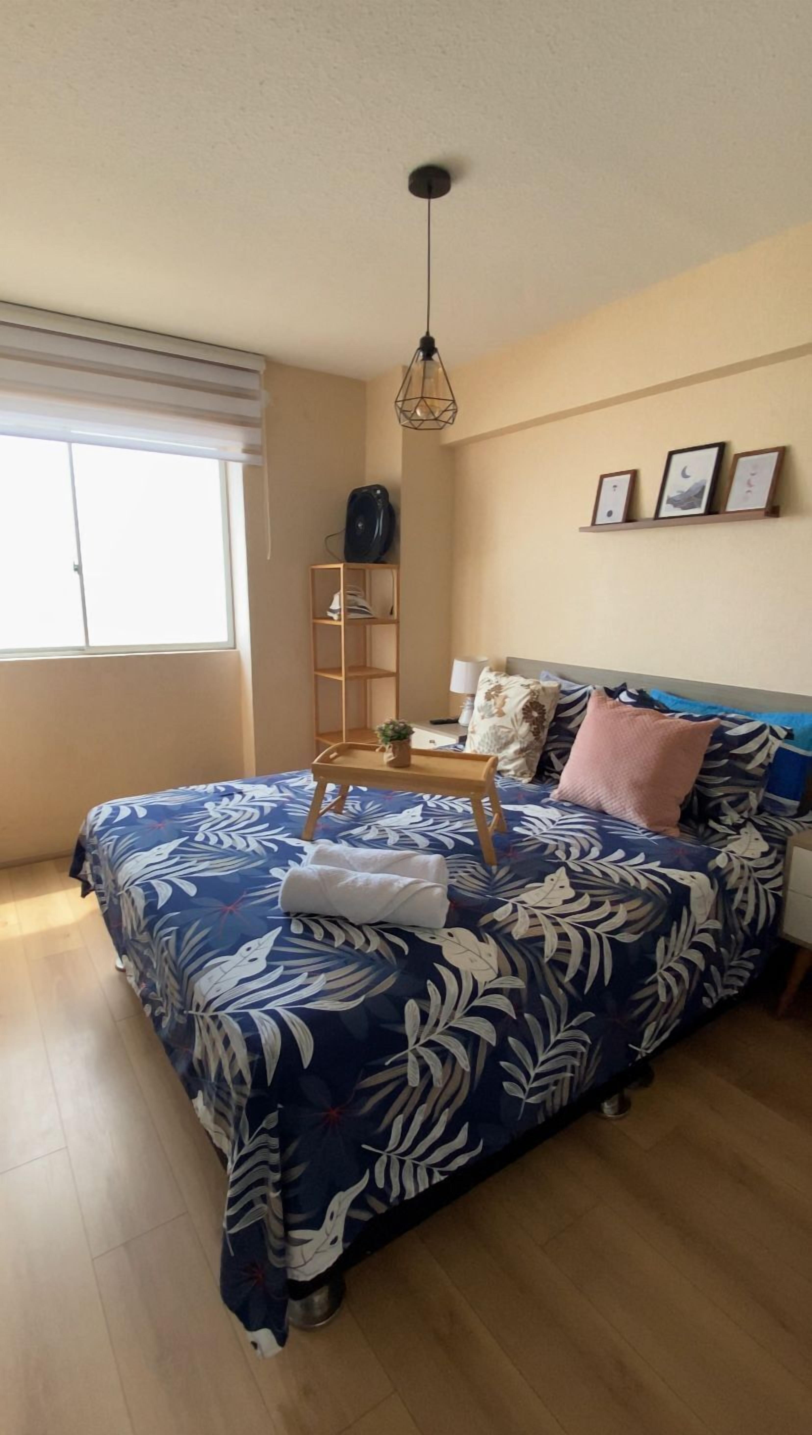 Un dormitorio con una cama, una mesita de noche, una lámpara, una estantería con libros y una ventana.