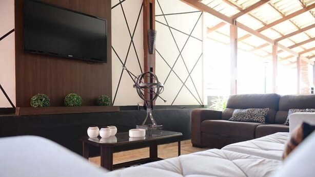 A modern living room with a flat-screen TV mounted on the wall, a wooden coffee table with decorative items, a sofa with patterned cushions, and a large window allowing natural light. - Ubaense Plaza Hotel (Uba)