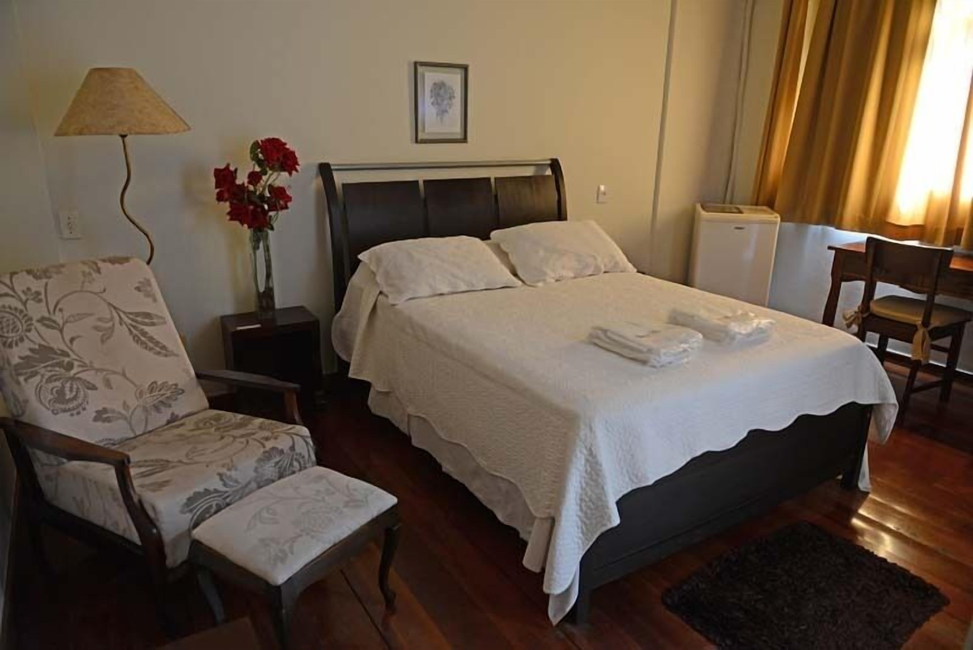 A bedroom with a bed, a chair, a nightstand, a lamp, a vase with flowers, and a picture on the wall.