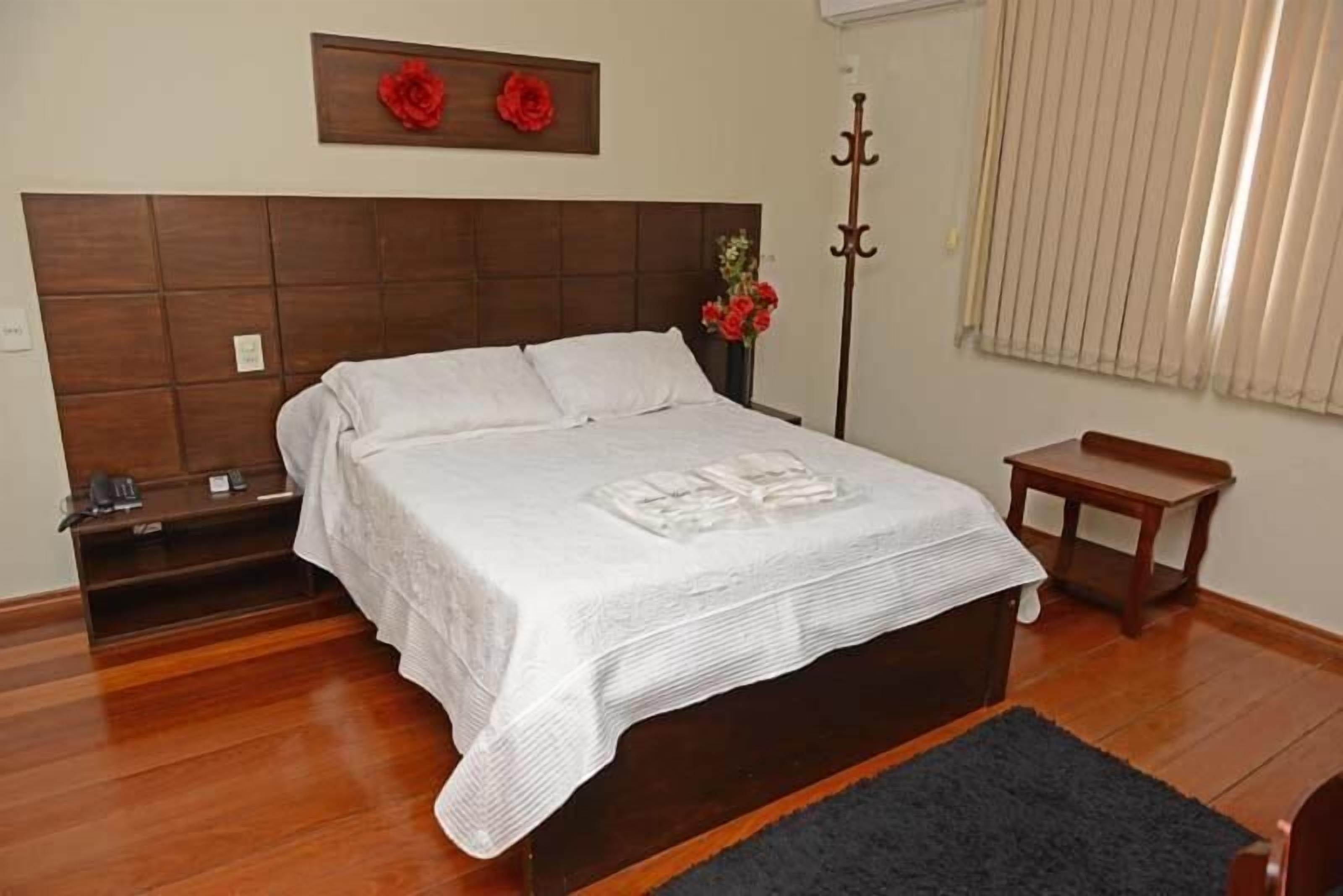 A neatly made bed with a white bedspread, a wooden headboard, a small bedside table, and a vase with red flowers.