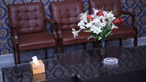 A room with three brown leather chairs, a glass table with a tissue box and remote control, and a vase with white and red flowers. - Family Hotel Lahore  (Block R 2)