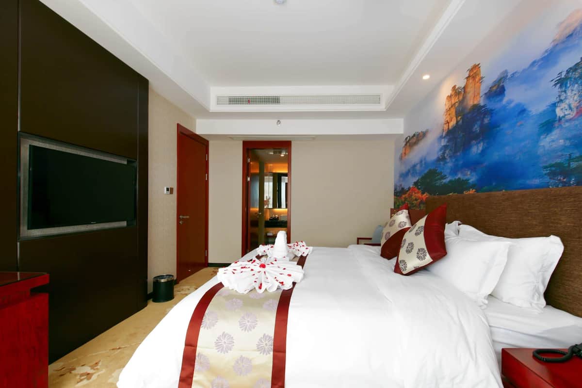 A hotel room with a large bed, a flat-screen TV, a painting on the wall, and a red side table.
