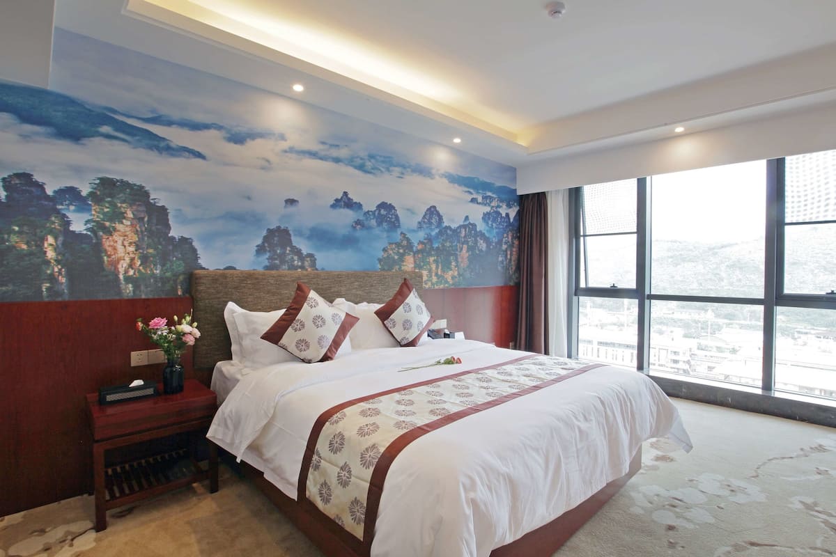 A hotel room with a large bed, a nightstand with a vase of flowers, a mural on the wall, and a large window offering a city view.