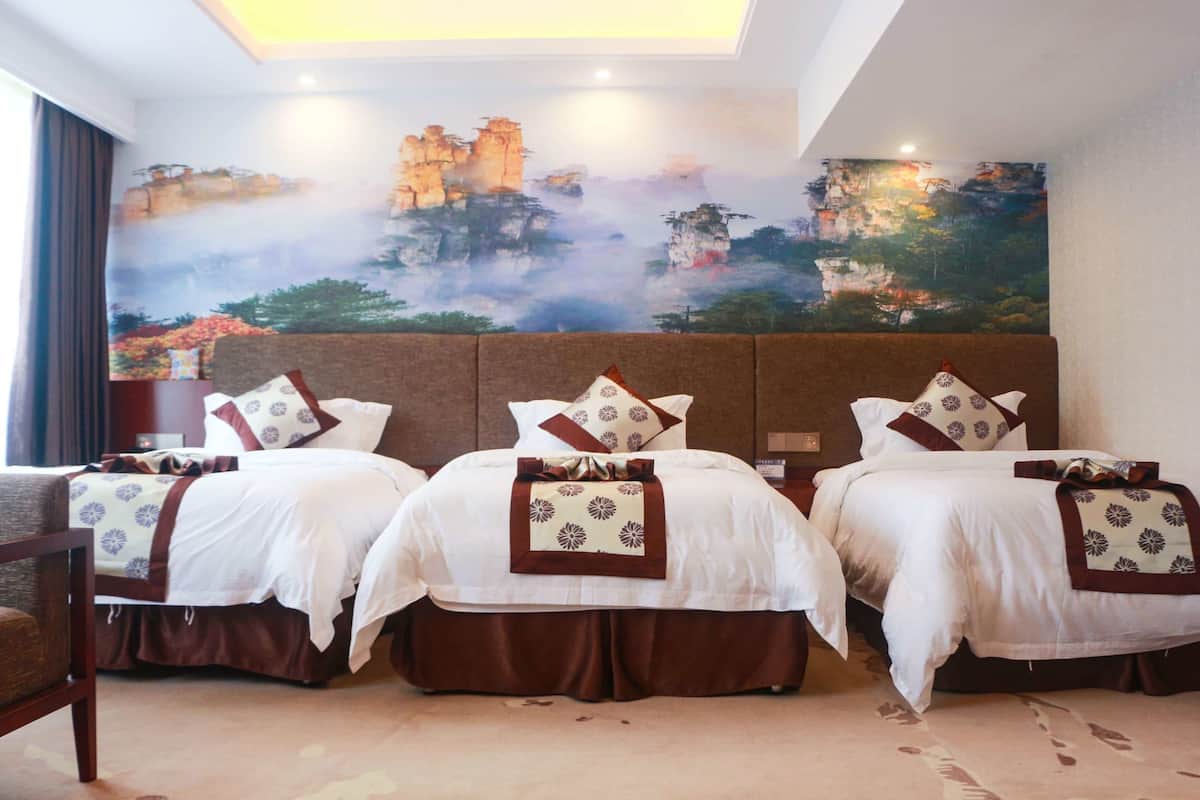 A hotel room with three beds, each with white bedding and brown accents, a large mural on the wall depicting a mountainous landscape, and a wooden chair.