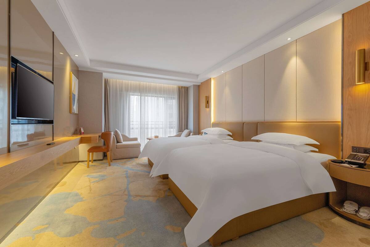 Ein Hotelzimmer mit zwei Betten, einem Schreibtisch, einem Fernseher und einem großen Fenster mit Vorhängen.