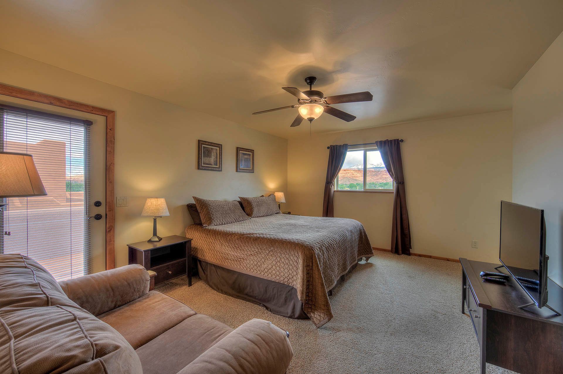 A bedroom with a bed, two bedside tables with lamps, a ceiling fan, a window with curtains, a TV, and a sofa.